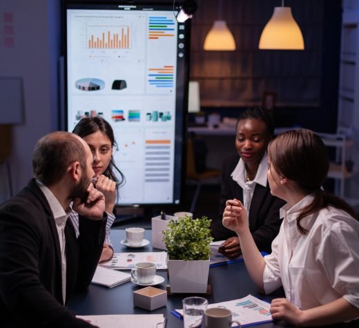 Workaholics businesspeople brainstorming financial company ideas analyzing strategy paperwork late at night in business office meeting room. Multi-ethnic coworkers checking solution